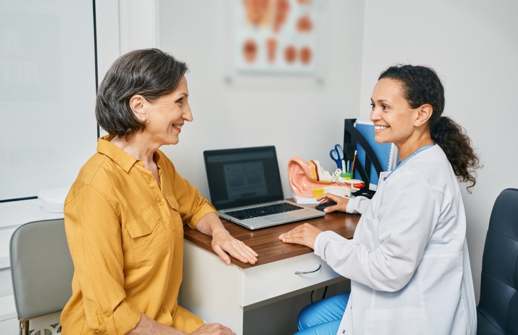 A smiling female audiologist treats an older woman wearing a mustard-colored shirt.