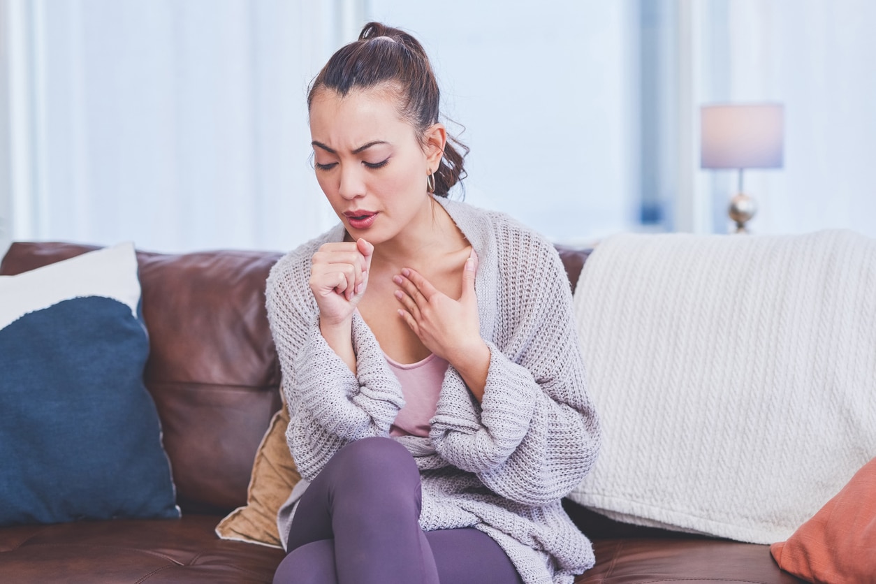 A girl experiencing a cough in a light purple sweater sitting on her couch.