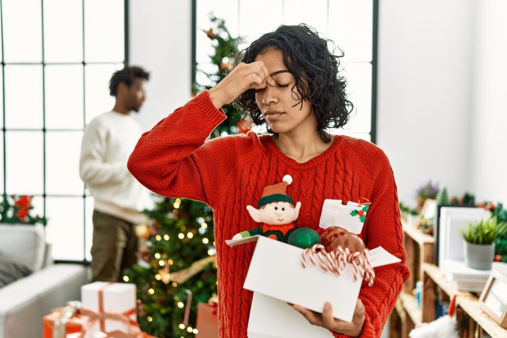 A woman standing by a Christmas tree with decorations tired, rubbing her nose and eyes, feeling nasal obstruction.