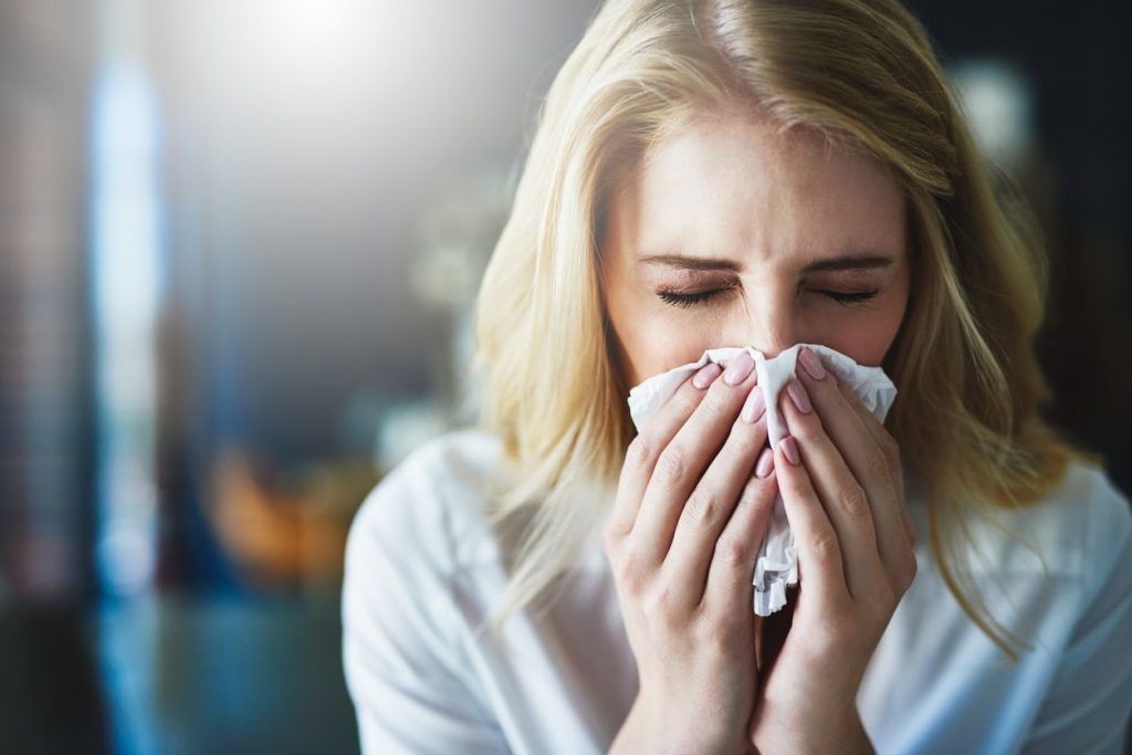 A blonde woman blowing her nose with the sunlight shining in the background.