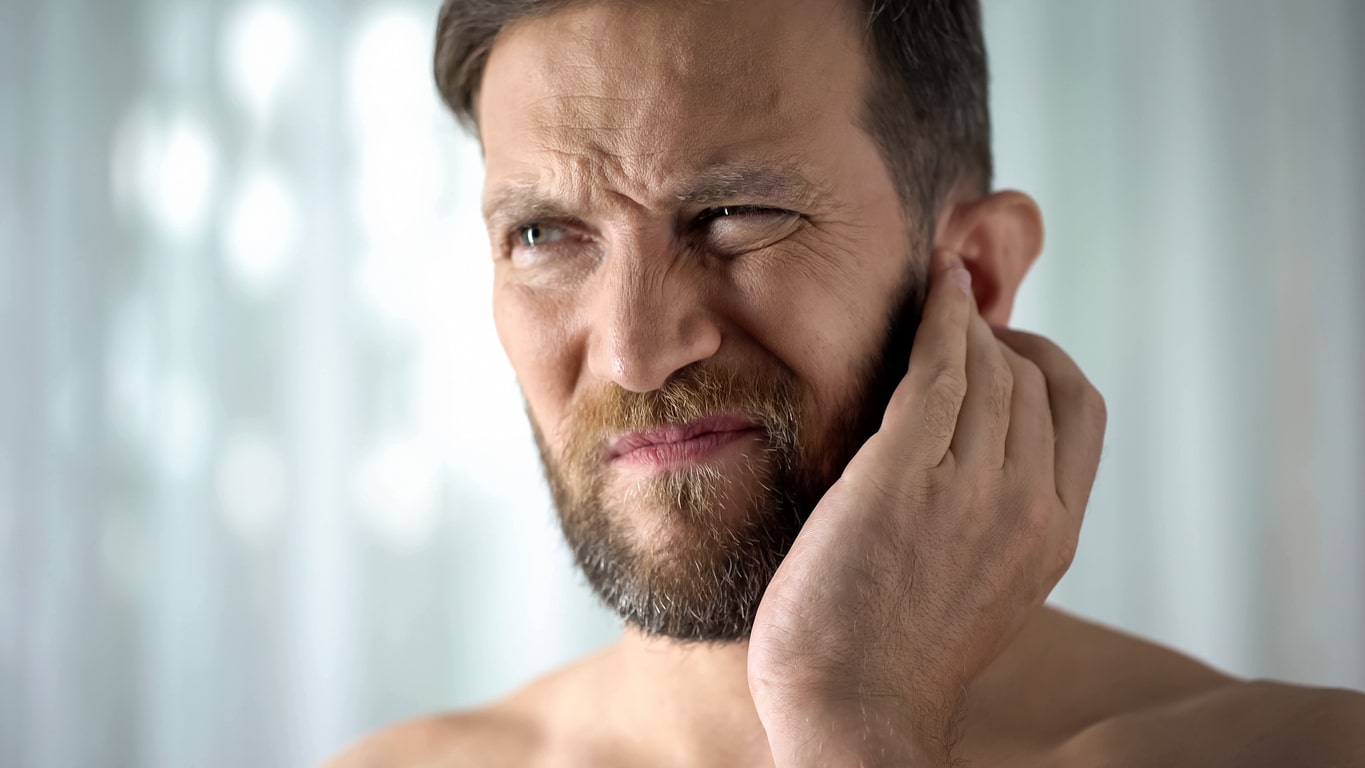 An adult man suffering holding his ear and grimacing in pain while suffering from an ear infection.