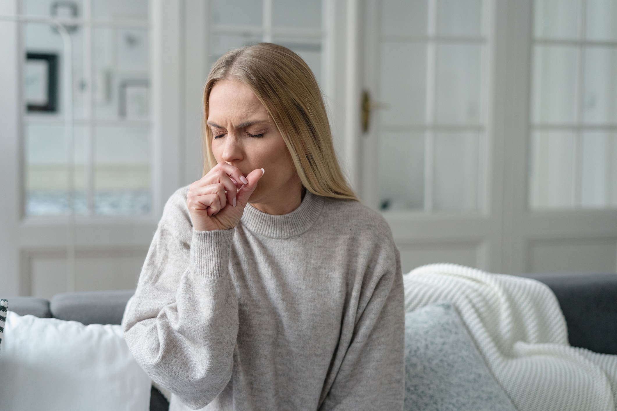 A woman sitting on a couch struggles with symptoms of irritable larynx syndrome.
