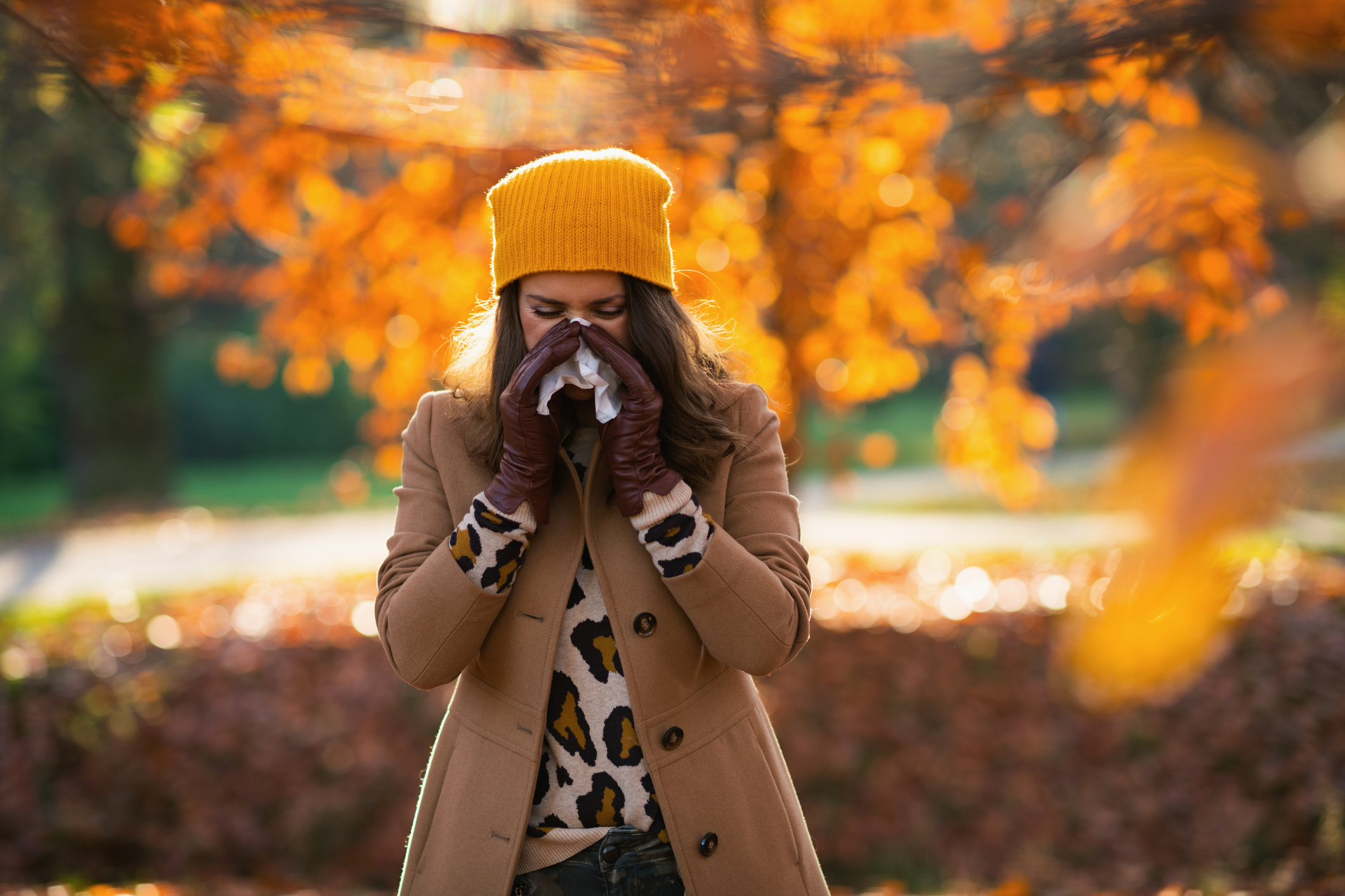 A woman wearing a yellow beanie blows her nose into a tissue while standing among autumn foliage, wondering if she has a cold or fall allergies.
